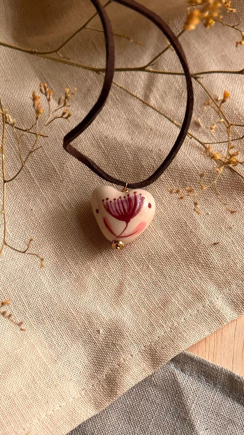 May include: A heart-shaped pendant necklace with a dark brown suede cord. The pendant is off-white with a hand-painted floral design in shades of pink and burgundy. A small gold-colored bead accents the bottom of the pendant.