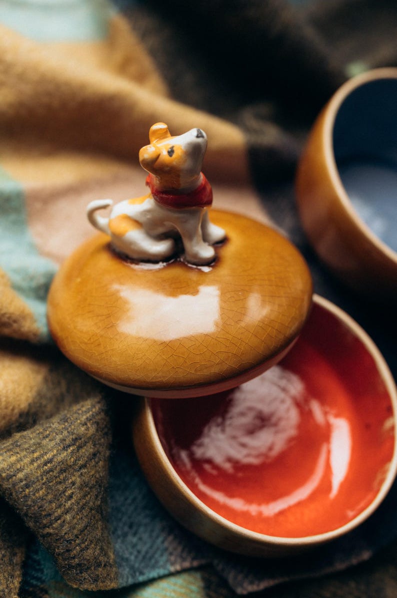 Handmade ceramic trinket box with warm amber crackle glaze lid topped by black and white spotted dog figurine, navy blue glossy base, autumn fall home decor, pet lover jewelry storage