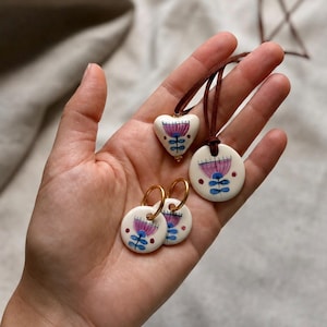 May include: A set of ceramic jewelry featuring a heart-shaped pendant, a circular pendant on a brown cord, and two circular hoop earrings. Each piece is white with a hand-painted floral design in shades of purple, blue, and red accents.
