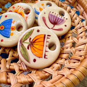 May include: A collection of off-white ceramic beads with hand-painted floral designs in orange, blue, purple, and green. The beads are round with a hole in the center and are displayed in a woven basket.