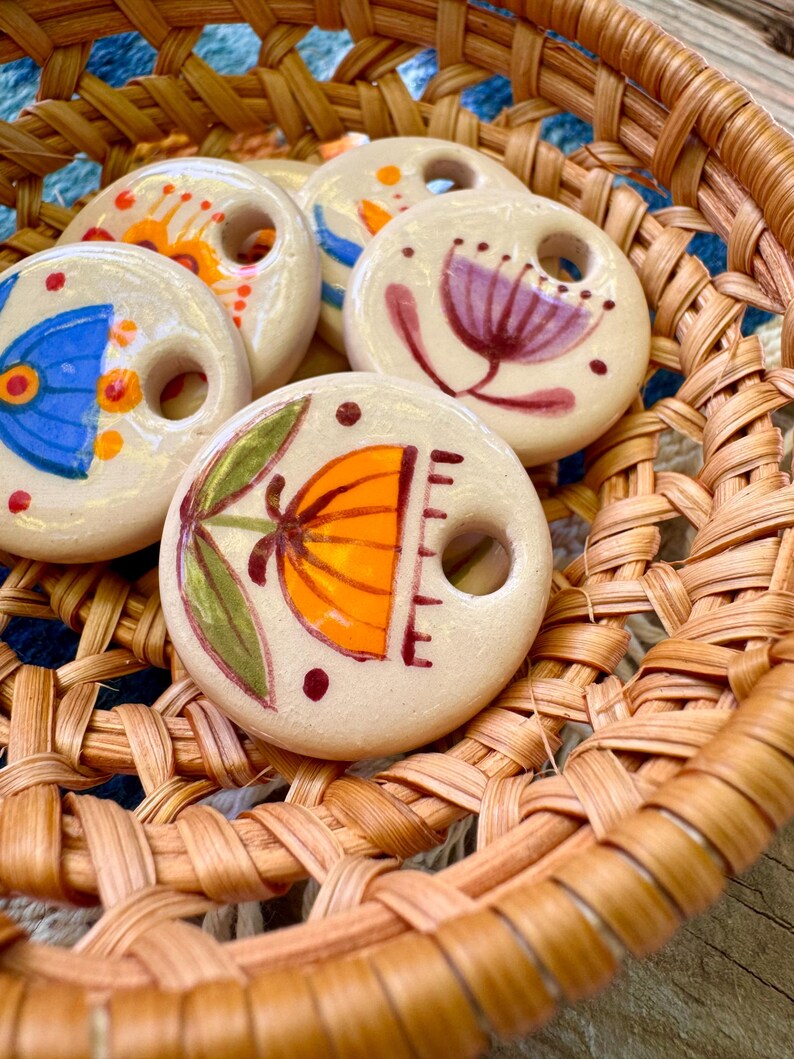 May include: A collection of off-white ceramic beads with hand-painted floral designs in orange, blue, purple, and green. The beads are round with a hole in the center and are displayed in a woven basket.
