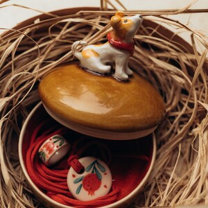 May include: A ceramic jewelry box with a brown lid featuring a small dog figurine. Inside, a red-lined compartment holds a ring and a pendant with a pomegranate design. The box is nestled in a woven basket.