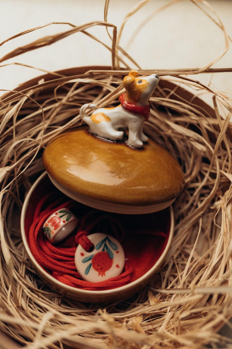 May include: A ceramic jewelry box with a brown lid featuring a small dog figurine. Inside, a red-lined compartment holds a ring and a pendant with a pomegranate design. The box is nestled in a woven basket.