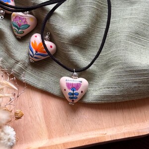 May include: Hand-painted heart-shaped pendants on black cords. Each pendant features a unique floral design in vibrant colors. The necklaces rest on a green fabric, with a wooden tray and brown fabric in the background.