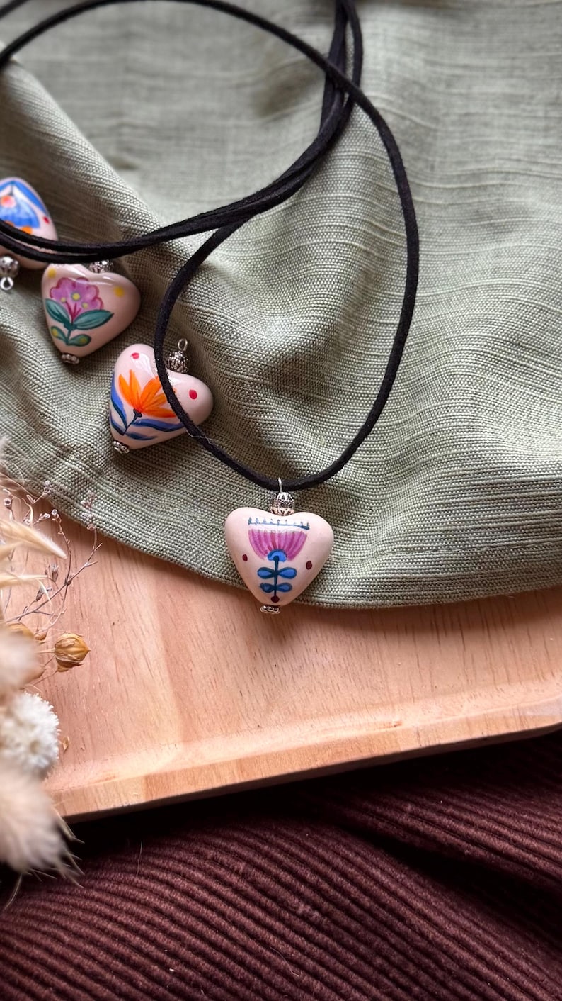 May include: Hand-painted heart-shaped pendants on black cords. Each pendant features a unique floral design in vibrant colors. The necklaces rest on a green fabric, with a wooden tray and brown fabric in the background.