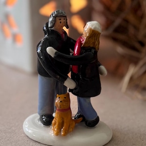 以下が含まれることがあります： 冬のコート、帽子、スカーフを着た、抱き合うカップルを描いた陶器製の置物。赤い首輪をした小さなオレンジ色の犬が足元にいます。置物は白い楕円形の台座にあります。