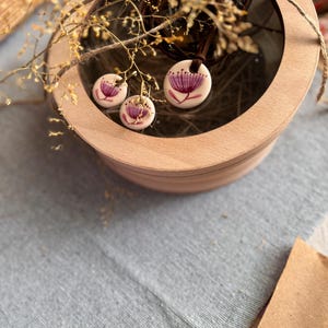 May include: A jewelry set featuring a pendant necklace and matching earrings. The white ceramic pieces are adorned with a purple floral design. The set is displayed inside a wooden box with a clear lid, and is accented with dried branches.