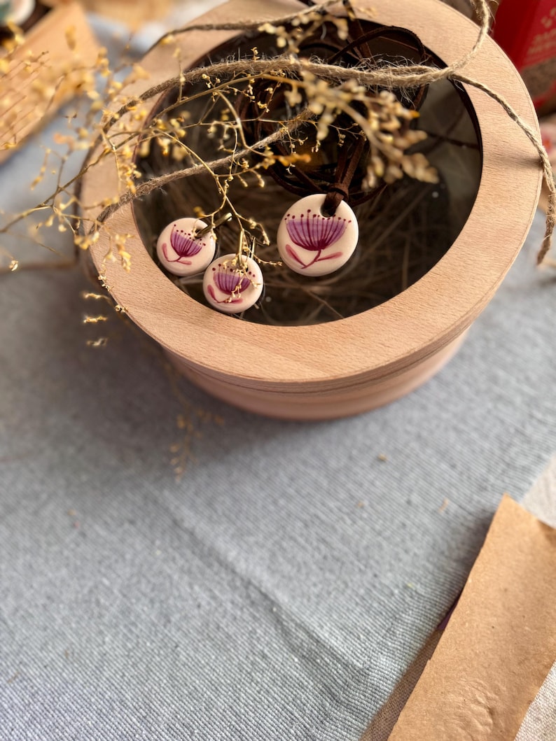 May include: A jewelry set featuring a pendant necklace and matching earrings. The white ceramic pieces are adorned with a purple floral design. The set is displayed inside a wooden box with a clear lid, and is accented with dried branches.