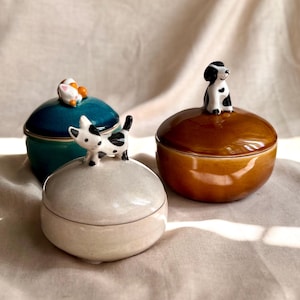 Puede incluir: Tres tarros de cerámica con tapa en color verde azulado, marrón y blanco roto. Cada tarro está coronado con una pequeña figura de animal: un gato, un perro y un perro blanco y negro. Los tarros están sobre un fondo de tela neutra.