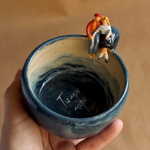 Può includere: Una ciotola in ceramica blu con un motivo a onde dipinto all'interno. Una piccola statuina di una coppia è appoggiata sul bordo. La ciotola ha la scritta "Ти мое море" all'interno.
