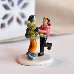 Può includere: Una statuetta in ceramica raffigura due pattinatori. Una figura indossa una giacca gialla e verde, pantaloni arancioni e pattini blu. L'altra figura indossa una giacca rosa e pantaloni neri. La statuetta è su una base bianca.