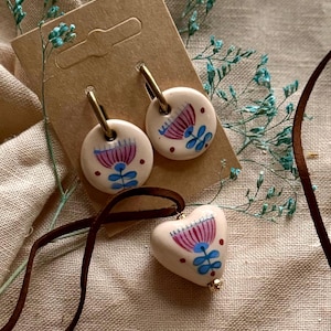 May include: A set of handmade ceramic jewelry featuring a floral design. The set includes a pair of round earrings with gold hoops and a heart-shaped pendant on a brown leather cord. The floral design is in pink, blue, and red.
