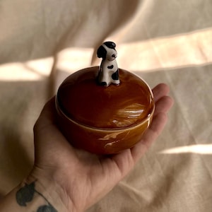 Könnte beinhalten: Eine braune Keramik-Schmuckdose mit Deckel, gekrönt von einer kleinen, schwarz-weißen Hundefigur. Die Dose ist rund und glänzend, gehalten in einer Hand vor neutralem Hintergrund.