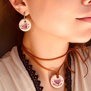 May include: A matching jewelry set featuring a floral design. The set includes a pair of round earrings and a pendant necklace with a brown leather cord. The floral design is pink and purple on a white background.