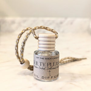 May include: A small glass bottle with a brown label that reads "DIP THE WICK CANDLE GUILTY PLEASURE Amber Orchid, Driftwood White OIL DIFFUSER". The bottle is hanging from a beige and gold cord with a wooden bead.