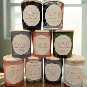May include: A collection of ten scented candles in glass jars with wooden lids. Each candle has a label with text such as "Night Shift Terrors" and "High Acuity Low Morale". The candles are arranged on a wooden surface.