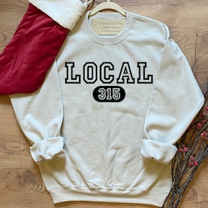 May include: A light gray sweatshirt with the text "LOCAL 315" in black block letters. The number 315 is in a black oval. A red Christmas stocking is in the upper left corner. The sweatshirt is on a wooden surface.