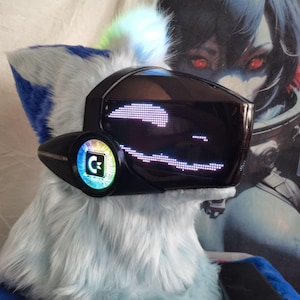 May include: A furry headpiece featuring light blue and white faux fur with a black visor displaying a white digital pixelated smile. The side of the visor includes a circular panel with a glowing, multicolored circuit board pattern and a letter C.
