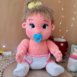 May include: A crocheted pink baby doll with blue eyes, a pacifier, and a white diaper. The doll is wearing white socks and has brown hair with a yellow crown.