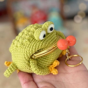 Pode incluir: Uma carteira de moedas de crochê verde feita à mão, em forma de sapo, com olhos e pés de contas laranja. O sapo tem um fecho de metal dourado e um porta-chaves dourado. Os olhos do sapo são brancos com pupilas pretas.