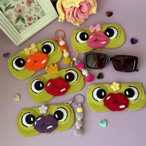 Peut inclure: Cinq étuis rectangulaires en crochet vert lime, chacun arborant de grands yeux blancs et noirs ainsi qu'une paire de lèvres proéminente orange, rose, rouge ou violette. Certains incluent des nœuds, des couronnes et des porte-clés perlés.