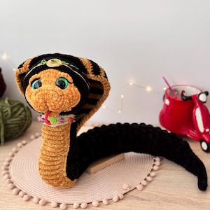 May include: A crocheted plush toy snake with a black and brown striped body, a golden head, and green eyes. The snake is wearing a gold crown and a colourful beaded necklace.