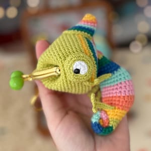 Puede incluir: Un camaleón de ganchillo hecho a mano en tonos verdes, azules, rosas y amarillos. El camaleón tiene un cierre dorado y una bolita verde en la punta de la lengua. La cola está rayada con múltiples colores.
