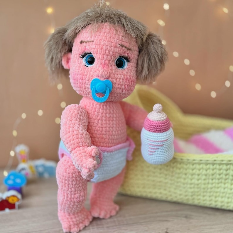 May include: A pink crocheted baby doll with brown hair, blue eyes, and a pacifier. The doll is holding a pink and white crocheted bottle. The doll is sitting in a yellow and white crocheted crib.