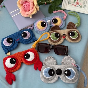 May include: A selection of colourful, handmade crochet eyeglass cases. The cases, in shades like blue and orange, feature large, cartoon-style eye designs. A pair of sunglasses is placed amongst the cases. Measurements are not applicable.