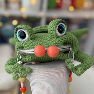 以下が含まれることがあります： 手作りの緑色のカギ針編みカエル型小銭入れ。大きな目とオレンジ色のビーズが特徴です。シルバーの留め金と、遊び心のあるデザインが施されています。小物を入れるのに最適です。