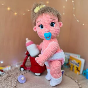 May include: A pink crocheted baby doll with brown hair, wearing a white diaper and white socks. The doll is holding a blue pacifier and is standing next to a red toy.