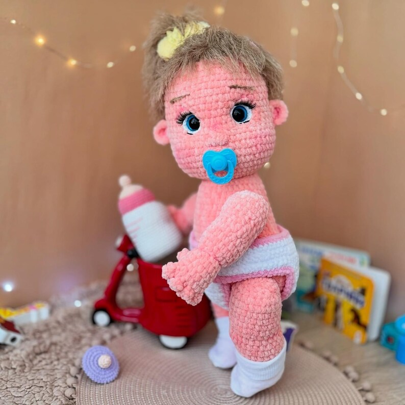 May include: A pink crocheted baby doll with brown hair, wearing a white diaper and white socks. The doll is holding a blue pacifier and is standing next to a red toy.