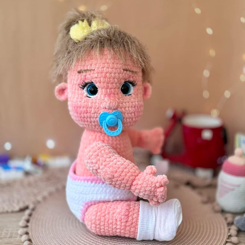 May include: A crocheted pink baby doll with a yellow hair bow, blue eyes, and a blue pacifier. The doll is wearing a white diaper with a pink trim and white socks.
