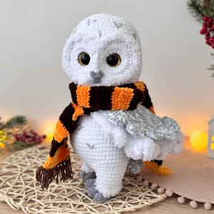 May include: White crocheted owl plush toy wearing an orange and brown striped scarf. The owl has large, black eyes with gold glitter accents.