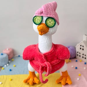 Pode incluir: Um ganso branco de crochê vestindo um roupão de banho rosa e uma toalha rosa na cabeça. O ganso tem fatias de pepino sobre os olhos.