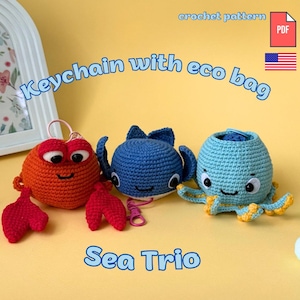Op de afbeelding: Drie gehaakte sleutelhangers van zeedieren, waaronder een rode krab, een blauwe walvis en een lichtblauwe octopus. De tekst luidt "crochet pattern", "Keychain with eco bag" en "Sea Trio". Een PDF-pictogram en een Amerikaanse vlag zijn zichtbaar.