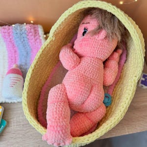 May include: A pink crocheted baby doll with brown hair lying in a yellow crocheted basket. The doll is wearing a blue pacifier.