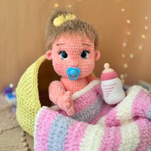 May include: A crocheted baby doll with pink skin, blue eyes, and a pacifier in its mouth. The doll is wrapped in a pink, white, and gray striped blanket and is sitting in a yellow crocheted basket. The doll is holding a small crocheted bottle.