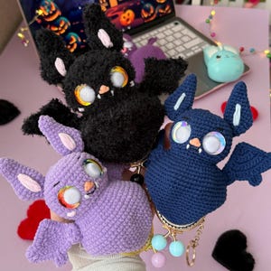 Könnte beinhalten: Drei gehäkelte fledermausförmige Beutel in Schwarz, Lavendel und Marineblau. Jede Fledermaus hat große schillernde Augen, kleine rosa Nasen, weiße Fangzähne und spitze Ohren. Der marineblaue Beutel besitzt einen goldfarbenen Metallrahmen und einen Schlüsselanhänger.