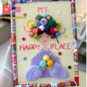 Puede incluir: Un tapiz de pared de ganchillo hecho a mano con una cara sonriente con pelo de hilo y las palabras "MY HAPPY PLACE". La obra de arte incluye bolas de hilo de colores, botones y una forma púrpura. El marco está decorado con papel.