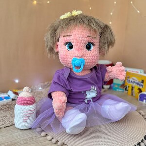 May include: A crocheted doll with pink yarn, blue eyes, and a purple shirt with a unicorn design. The doll is wearing a purple tutu and has a blue pacifier in its mouth. The doll is sitting on a brown and white rug.