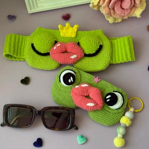 Puede incluir: Una diadema de ganchillo verde con una corona amarilla y labios rosas, junto a un estuche de ganchillo verde con ojos grandes, labios rosas y una pequeña corona rosa, acompañado de gafas de sol de montura oscura y un llavero de cuentas amarillas.