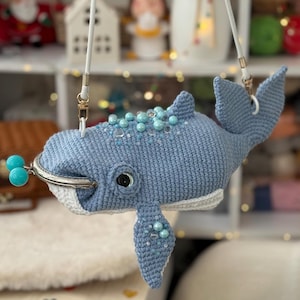 Op de afbeelding: Een lichtblauwe gehaakte walvisvormige tas met een witte riem. De walvis heeft een zilveren sluiting, een zwart oog en is versierd met blauwe kralen. Een uniek accessoire.