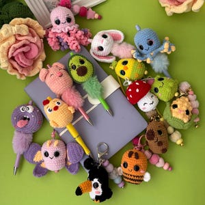 May include: A collection of colorful crochet animal and food-themed pens and keychains featuring a pink octopus, a white bunny, a blue octopus, a pig, an avocado, a mushroom, a donut, a chick, a butterfly, a toucan, a bee, and a corn cob.