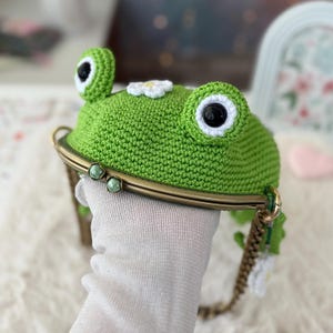 以下が含まれることがあります： 緑色のカギ針編みで作られたカエルの形をした小銭入れ。大きな丸い目と小さな白い花が付いています。小銭入れはブロンズ色の金属フレームとチェーンストラップが付いています。カエルの目は黒と白です。