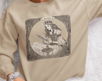 Puede incluir: Una sudadera beige con un estampado gráfico de una mujer leyendo un libro. El gráfico es en blanco y negro y presenta a una mujer con una corona en la cabeza. El texto "The Reader" está por encima del gráfico.