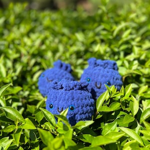May include: Three handmade, plush, blue crocheted creatures with smiling faces and large, round eyes. The soft toys are nestled among vibrant green leaves, creating a playful and whimsical scene. The toys are a deep shade of blue.
