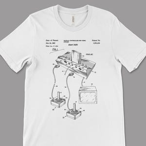 Op de afbeelding: Wit t-shirt met een zwart-wit diagram van een Atari 2600 videogameconsole en controllers. Het diagram bevat de tekst "Manual Controller for Video System Atari 2600" en patentinformatie.