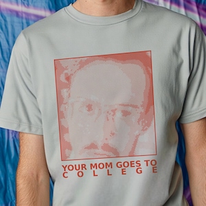 May include: A light gray t-shirt featuring a red-toned portrait and the text "YOUR MOM GOES TO COLLEGE" in red. The graphic is a halftone image of a man's face. The shirt has short sleeves and a crew neck.