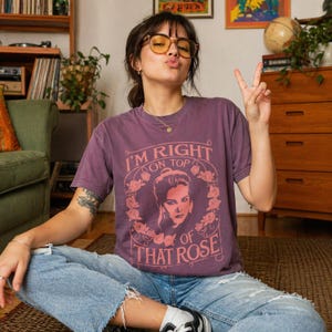 May include: A purple t-shirt with the text "I'M RIGHT ON TOP OF THAT ROSE" in a circular design with a woman's portrait and floral accents. The shirt is worn with blue jeans.